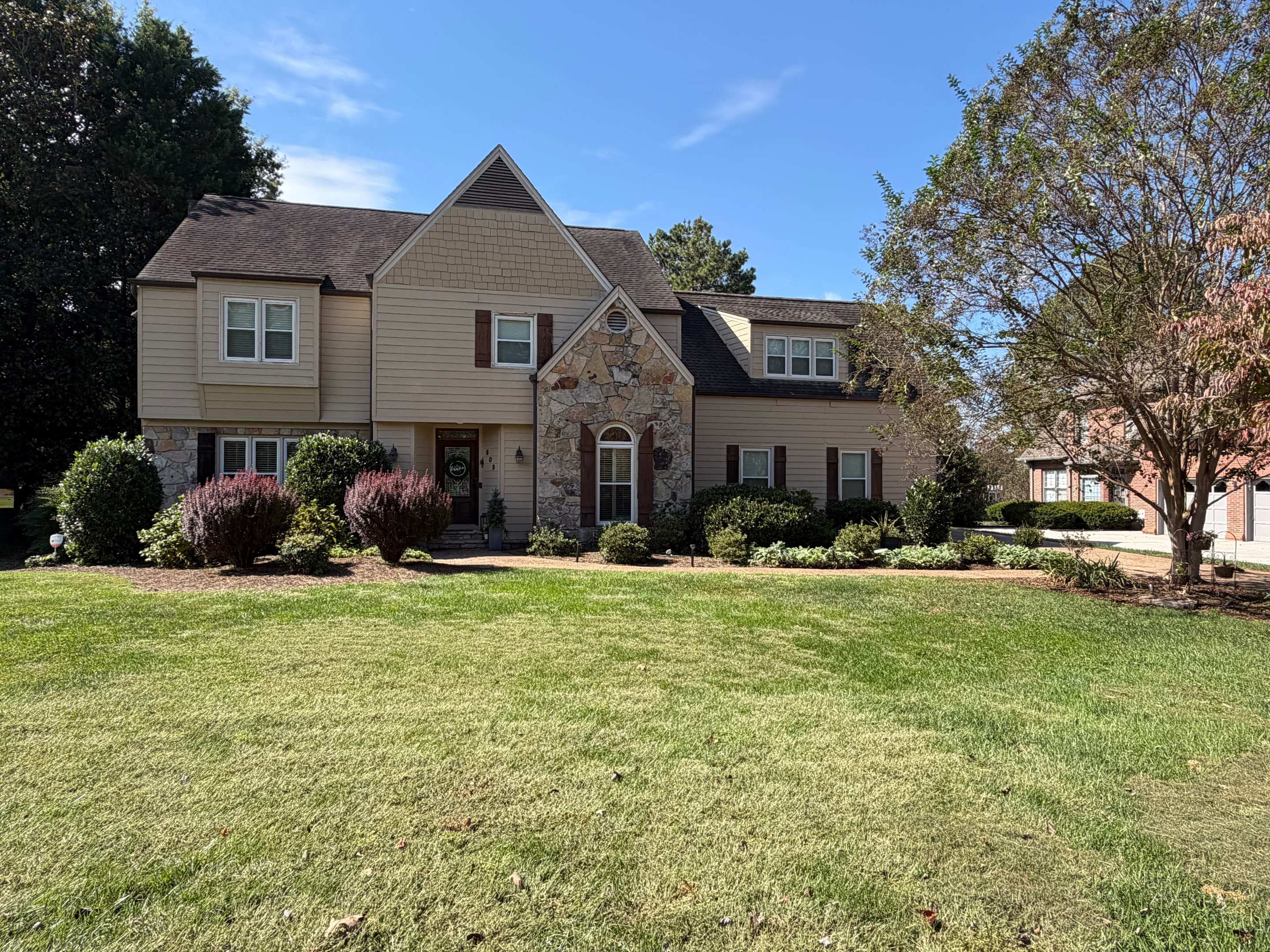 Two-story home with tan siding, a stone entry gable, and dark shutters behind a wide front lawn.